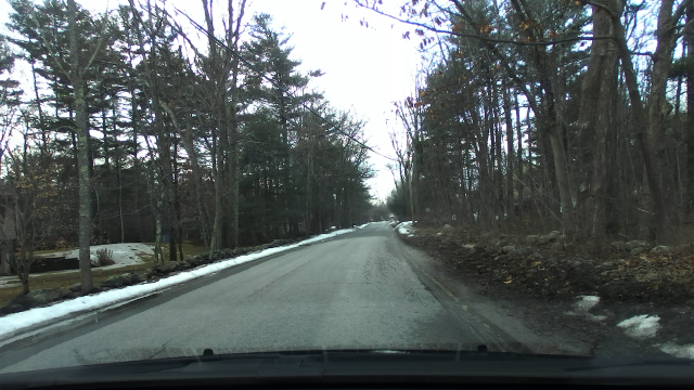 A largely straight unmarked road through a lightly wooded area with a clearing to the immediate left on a cloudy afternoon with some snow on the edges of edges of the road where it was mounded from snowplows. Most of the unpacked snow has melted leaving some wet areas on the road surface.