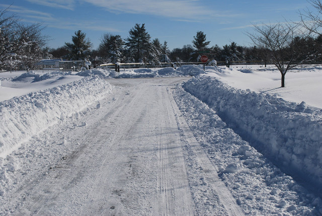 A picture of a two lane over flat terrain with about a foot of snow plowed cleanly to the edges of the road as to form very clear boundaries of the edges but not so cleanly as to be a sharp edge as if cut by a saw. The road surface is still entirely covered by a thin layer of compacted snow thick enough to be unable to see the color or texture of the road surface and remaining white enough to be similar in color to the snow banks at the edge of the road. A stop sign and intersection is about 10m in front of the camera.